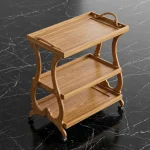 Sheesham wood kitchen trolley with three shelves, curved S-shaped sides, small handles, and black wheels on black marble floor by Haq Bahoo Furniture